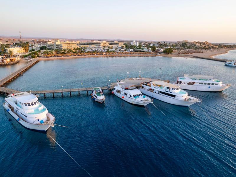 Cinq yachts amarrés à un quai dans une eau bleue calme près d'une ville côtière au coucher du soleil.