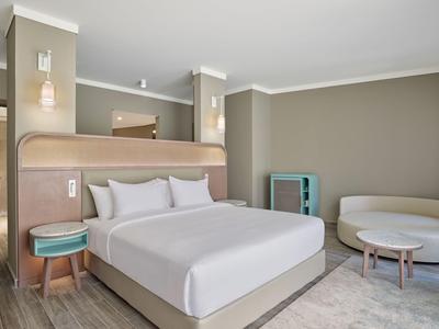 Moderní hotelový pokoj s manželskou postelí, nočními stolky, pohovkou a kulatým stolem v neutrálních tónech.