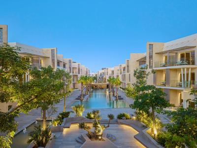 Moderní hotelový komplex s bazénem a zelení pod jasnou modrou oblohou.