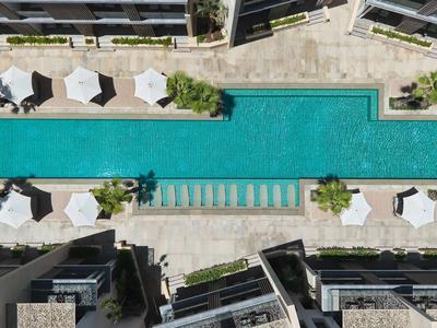Pool with clear water and white umbrellas between modern hotel buildings.