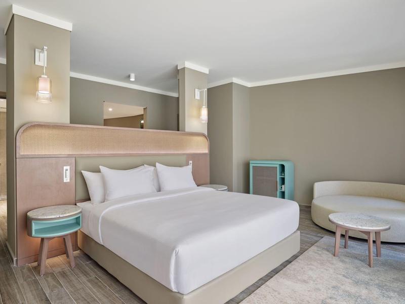 Chambre d'hôtel moderne avec lit double, deux tables de chevet, canapé et petite table aux tons neutres.