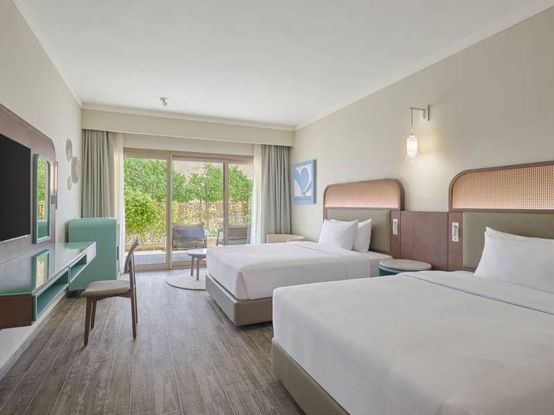 Chambre d'hôtel lumineuse avec deux lits, bureau, TV et terrasse donnant sur un jardin.