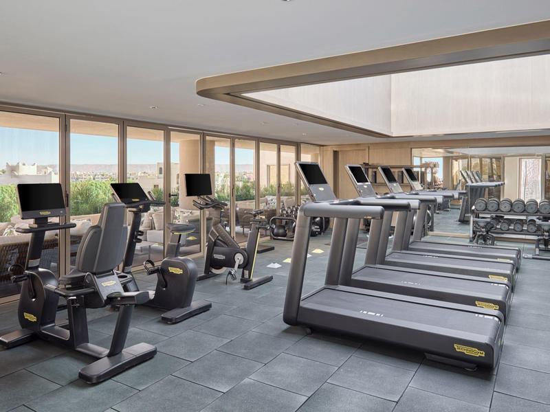 Salle de sport lumineuse avec tapis roulants, vélos et grandes fenêtres donnant sur l'extérieur.