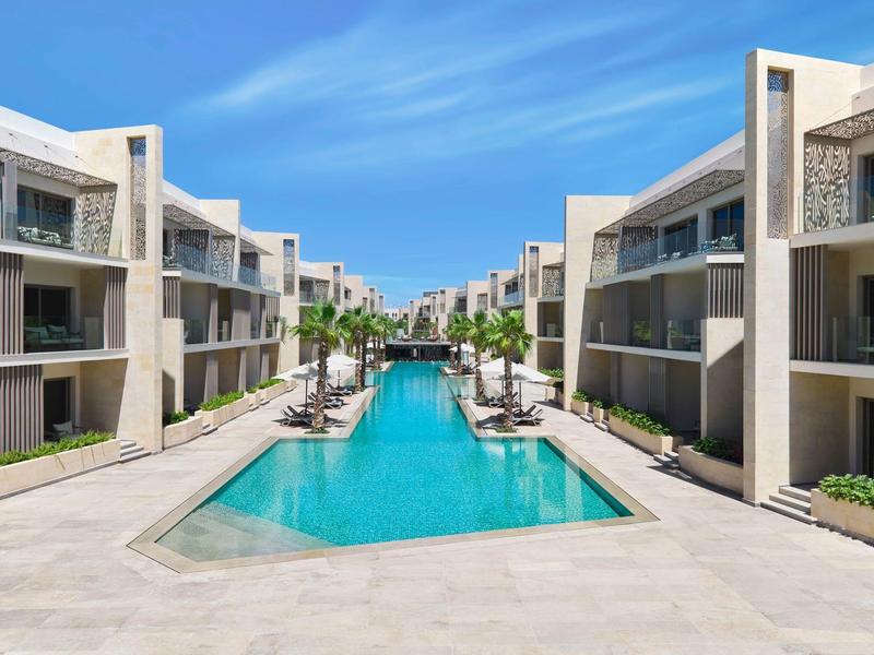Complexe hôtelier moderne avec une longue piscine, entourée de bâtiments de trois étages et de parasols.