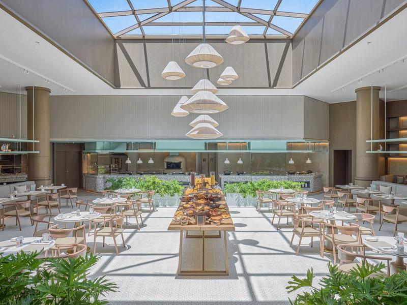 Restaurant moderne et lumineux avec des tables disposées au centre sous un toit en verre et des plantes vertes.