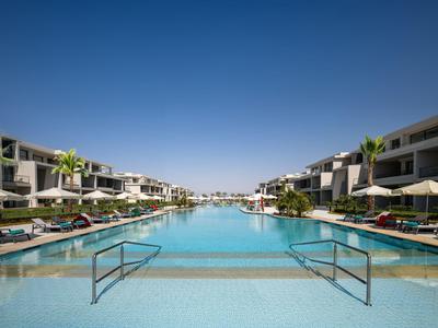 A large outdoor pool with lounge chairs and umbrellas next to modern hotel buildings under a clear blue sky.