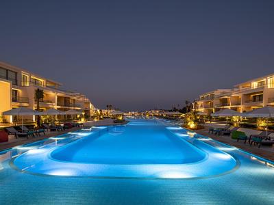Large illuminated hotel swimming pool with lounge chairs and buildings at night.