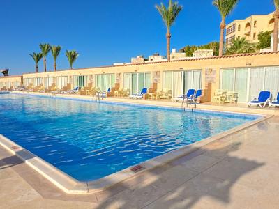 Grande piscine avec chaises longues devant un hôtel sous un ciel bleu clair.