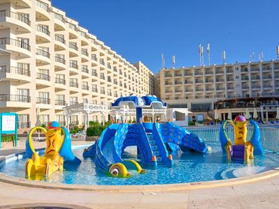 Zona de juegos acuática para niños frente a un hotel de varios pisos bajo un cielo despejado.