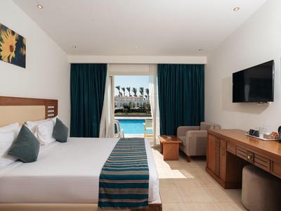 Habitación de hotel con cama doble, TV de pantalla plana y vista a la piscina bajo un cielo claro.