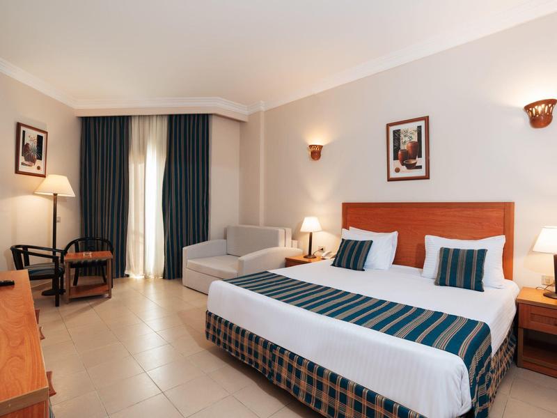 Habitación de hotel con cama doble, sofá, escritorio y decoración sencilla en tonos claros.