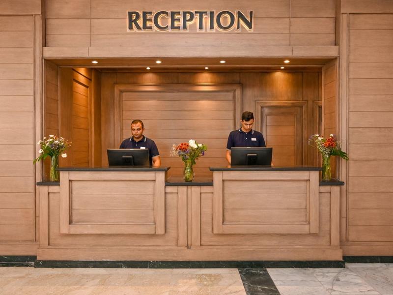 Recepción de hotel con dos recepcionistas detrás de mostradores de madera y arreglos florales.