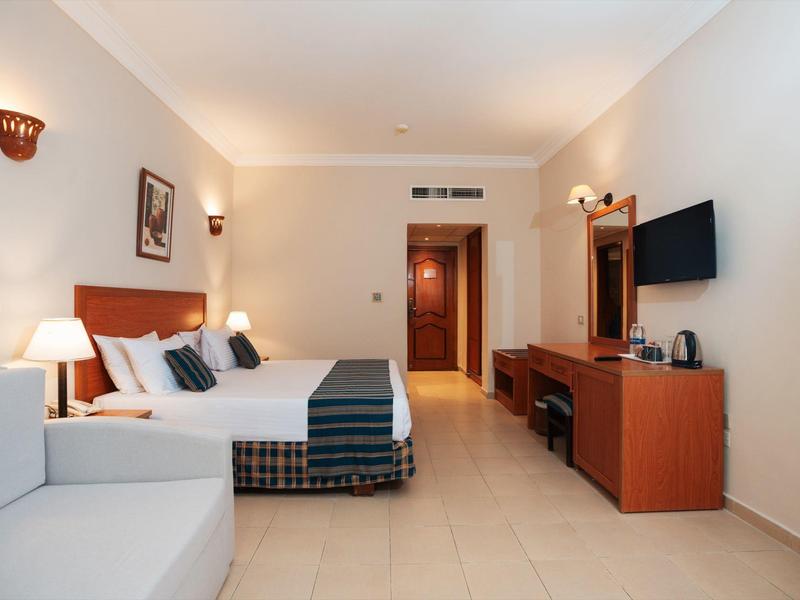 Habitación de hotel luminosa con cama doble, sofá, TV y muebles de madera sobre suelo de baldosas beige.