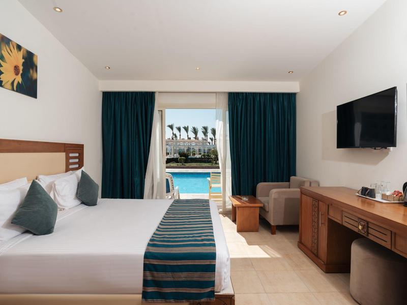 Habitación de hotel con cama doble, TV de pantalla plana y vista a la piscina bajo un cielo claro.