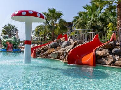 Vodní park s červenou skluzavkou a houbovým sprchou v bazénu obklopeném palmami.