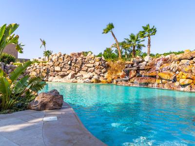 Modrý bazén s vodopádem a palmami pod jasnou oblohou v tropickém prostředí.