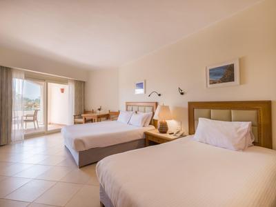 Světý hotelový pokoj se dvěma postelemi, nočním stolkem a výhledem na balkon se zahradou.