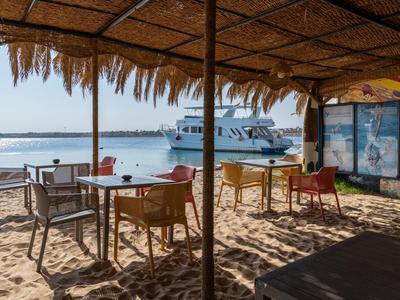 Slunečná plážová kavárna se židlemi a stoly, výhled na loď na vodě.