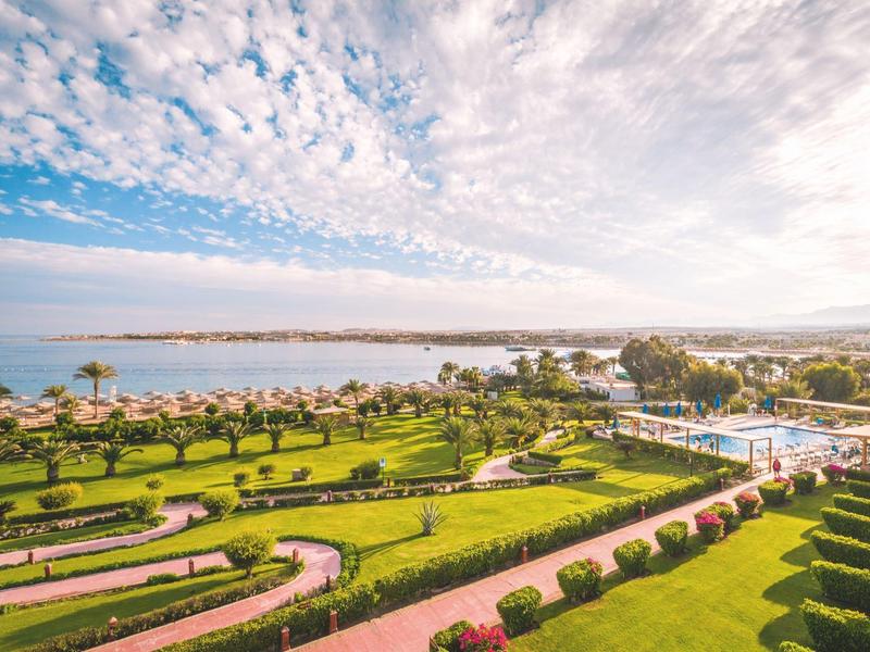 Grand jardin d'hôtel avec palmiers et vue sur la mer sous un ciel partiellement nuageux.