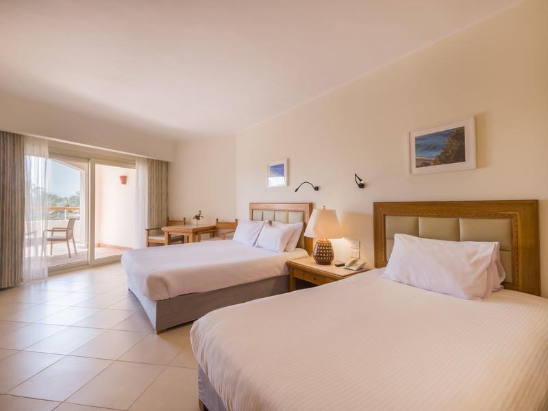 Chambre d'hôtel lumineuse avec deux lits, une chaise, un bureau et un balcon donnant sur l'eau.