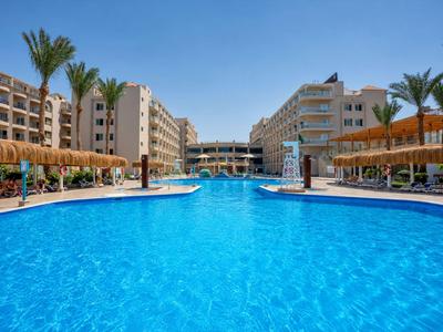 Large hotel pool with blue water, surrounded by palm trees and umbrellas under clear skies.