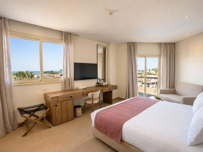 Bright hotel room with double bed, desk, chair, and sea view.