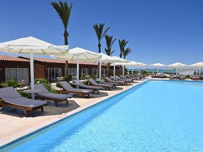 Strefa basenowa hotelu z leżakami pod białymi parasolami i palmami pod czystym niebem.