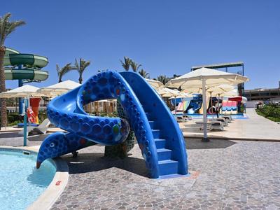Aire de jeux toboggan bleu à côté de la piscine avec parasols et palmiers en arrière-plan.