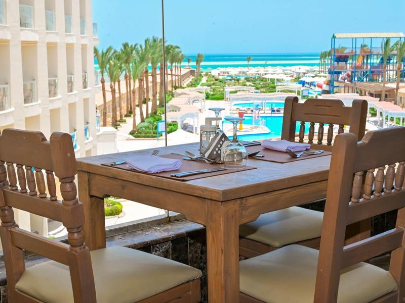 Table en bois avec quatre chaises sur un balcon donnant sur une piscine et la mer dans un hôtel.