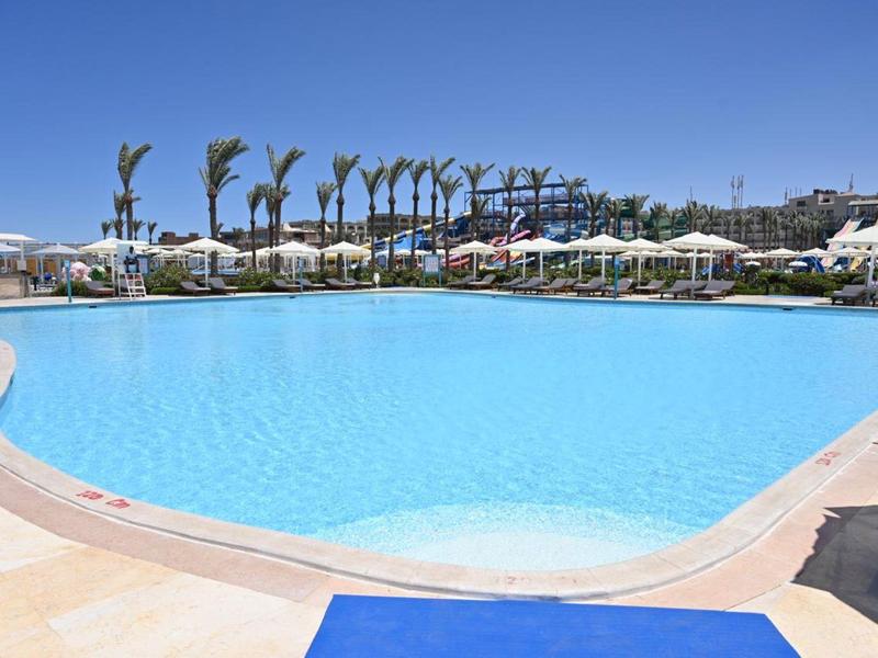Grande piscine avec eau bleu clair, transats et palmiers par temps ensoleillé.