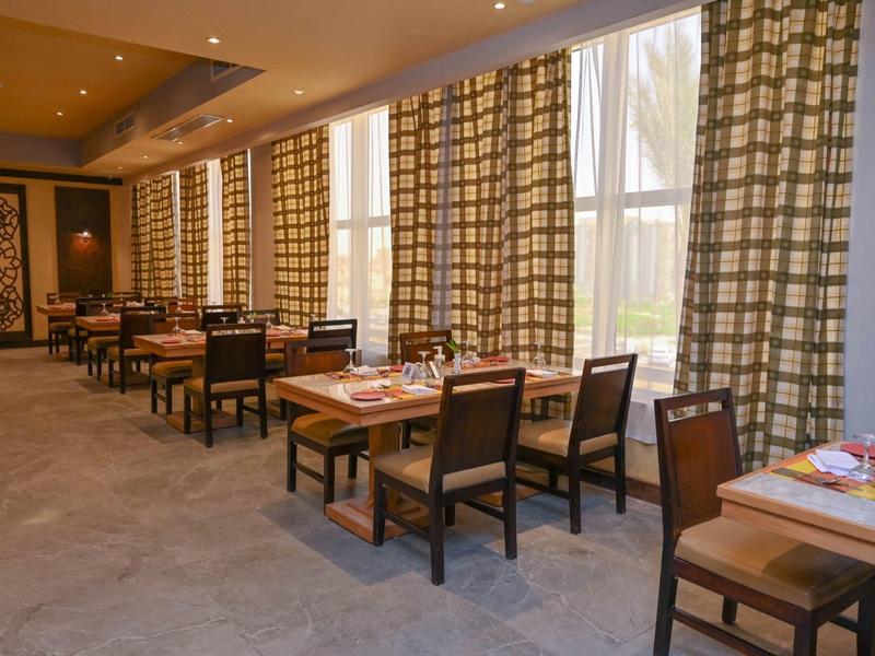 Salle à manger lumineuse avec tables en bois, chaises et grandes fenêtres avec rideaux à motifs.