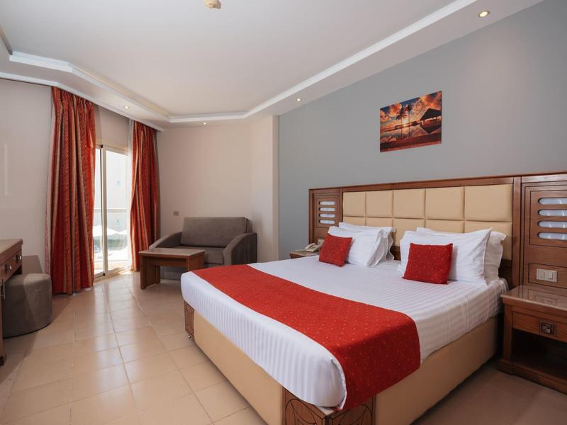 Camera d'hotel luminosa con letto matrimoniale, accenti rossi e divano vicino alla finestra con tende rosse