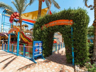 Entrée d'un parc aquatique avec des toboggans, des palmiers et des voiles d'ombrage sous un ciel clair.