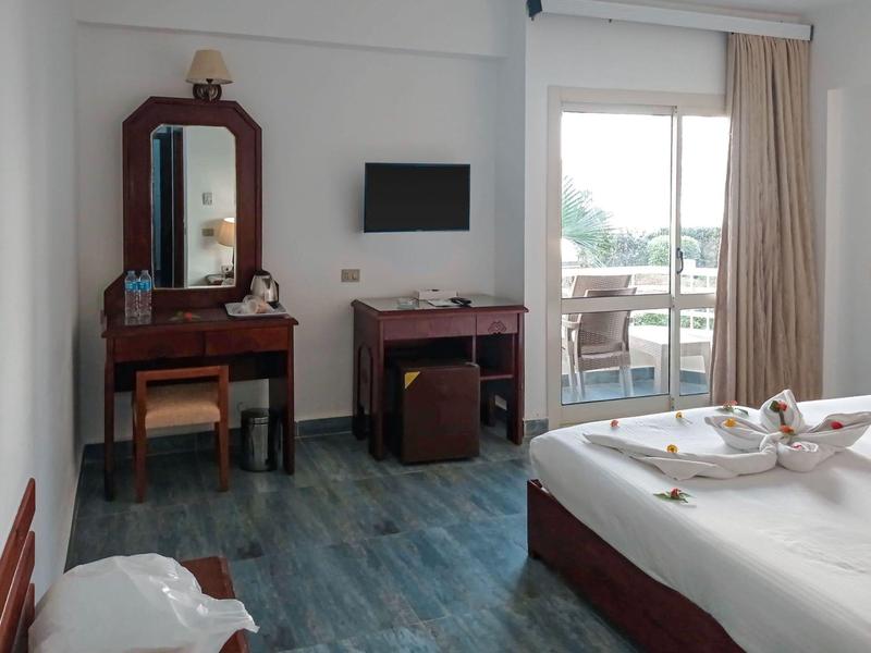 Chambre d'hôtel avec bureau, miroir, télévision, minibar et balcon avec rideaux.