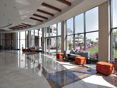 Světlá hotelová lobby s velkými okny, oranžovým posezením a lesklou mramorovou podlahou.