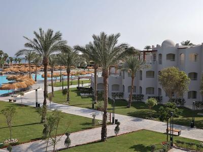 Hotelový komplex s bazénem, palmami a zelenými trávníky pod jasnou oblohou.