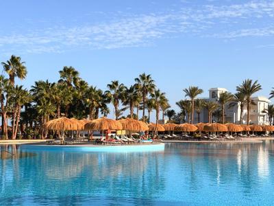 A calm pool with clear blue water in front of palm trees and umbrellas under a bright sky.