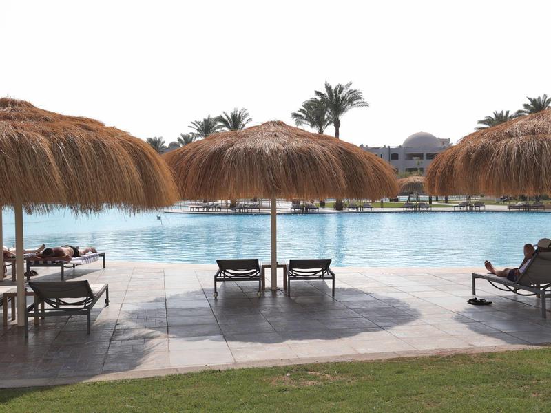 Plusieurs chaises longues sous des parasols en paille à côté d'une grande piscine extérieure par temps ensoleillé.