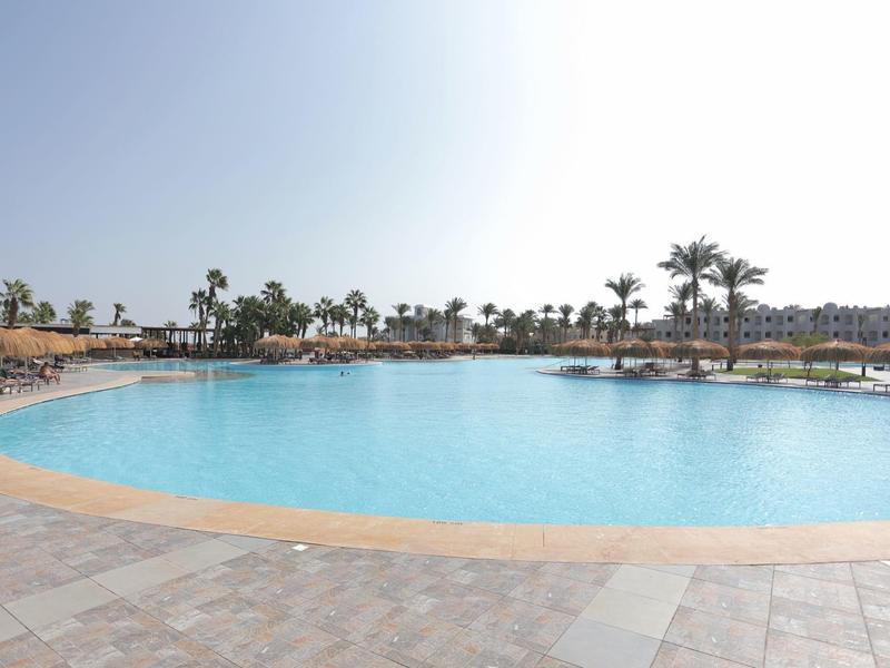 Grande piscine d'hôtel avec eau claire, entourée de palmiers et de chaises longues sous un ciel ensoleillé.