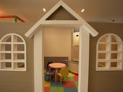 Casetta gioco per bambini con finestre e porta aperta in uno spazio interno con pavimento colorato.