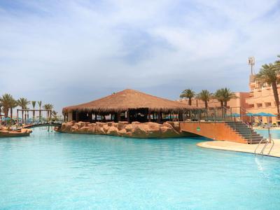 Großer Pool mit Sonnenterrasse und Strohüberdachtem Restaurant in einer Ferienanlage bei bewölktem Himmel.