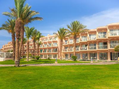 Hotelgebäude mit mehreren Etagen, Balkonzimmern und Palmen vor einem großen Rasen.