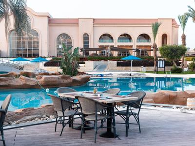 Außerhalb gedeckter Sitzbereich mit Tischen und Stühlen neben einem Pool und Hotelgebäude im Hintergrund.