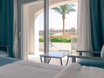 Chambre d'hôtel avec vue sur la terrasse, la piscine et les palmiers sous un ciel bleu.