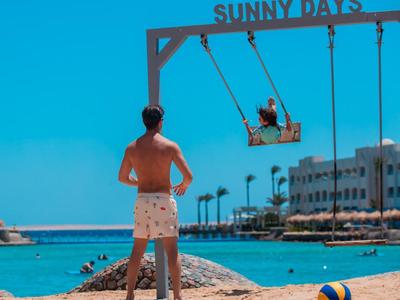 Homme et enfant sur une balançoire au bord de la mer sous un ciel bleu avec le texte 'SUNNY DAYS'.