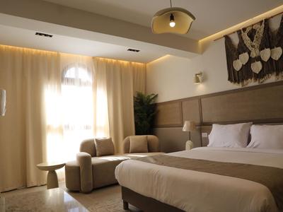Bright hotel room with large bed, two armchairs, curtains, and modern decor.