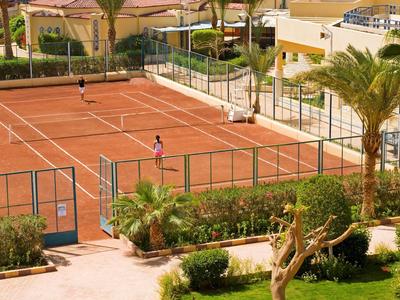 A red clay tennis court with two players, surrounded by garden and buildings.