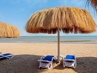 Pláž s pískem, dvěma lehátky a slaměnými slunečníky u vody.