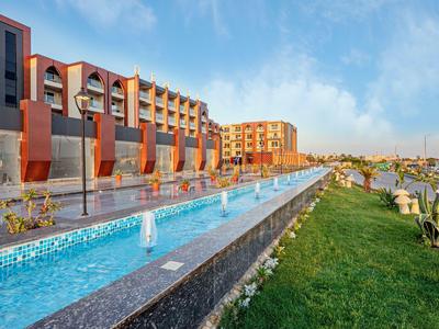 Moderní hotel s dlouhým vodním prvkem, fontánami a udržovanou trávou pod jasnou oblohou.