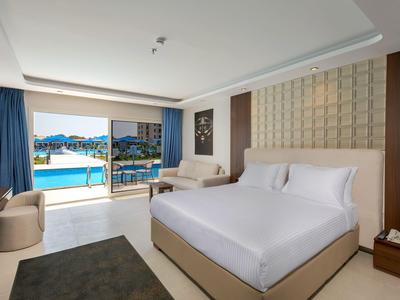 Moderní hotelový pokoj s velkou postelí, balkonem a výhledem na bazén za denního světla.